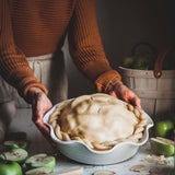 Person holding a raw apple pie in a white ceramic pie dish with apples around