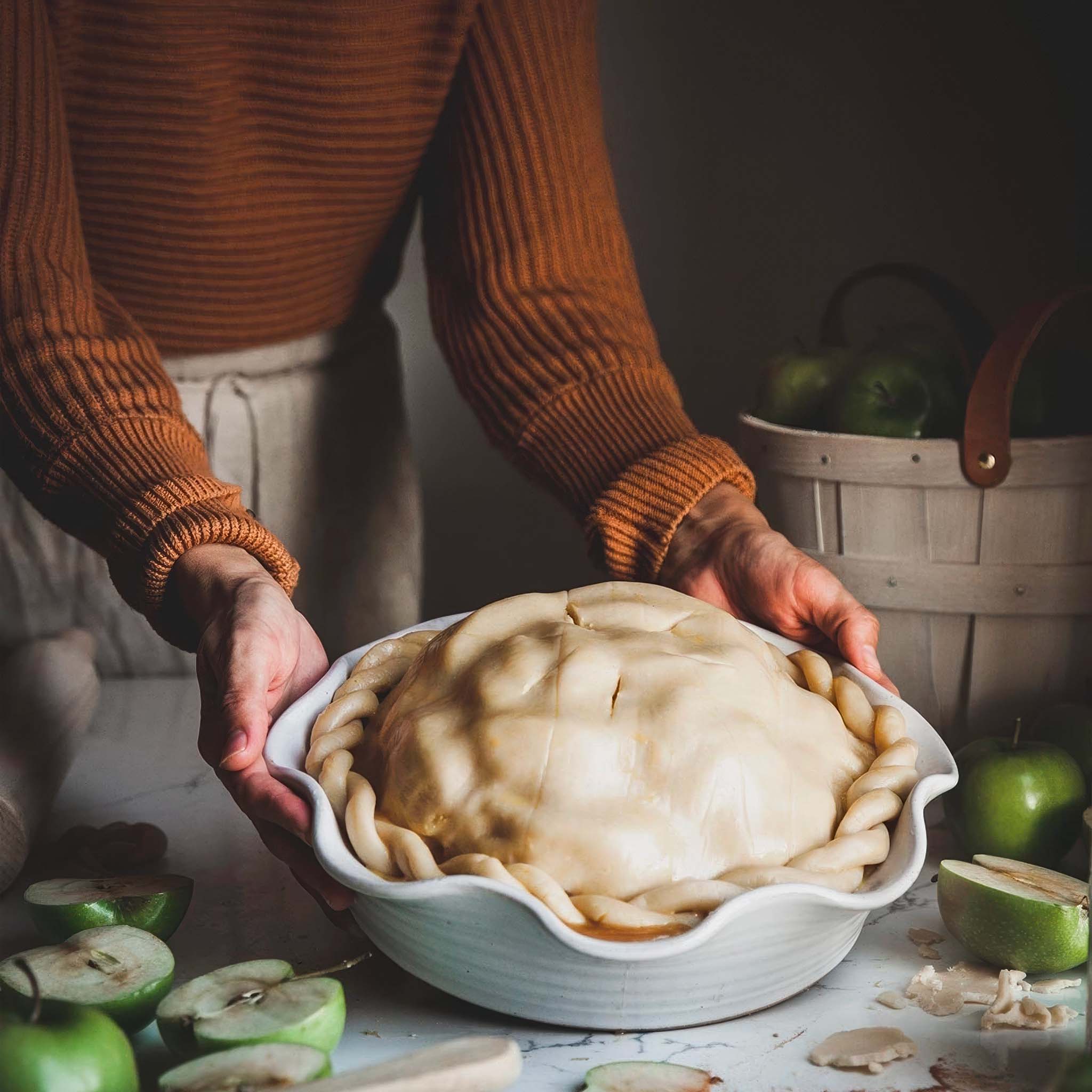 Person holding a raw apple pie in a white ceramic pie dish with apples around