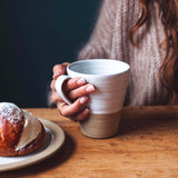 Person holding a white mug with a brown base on a wooden table next to a pastry.