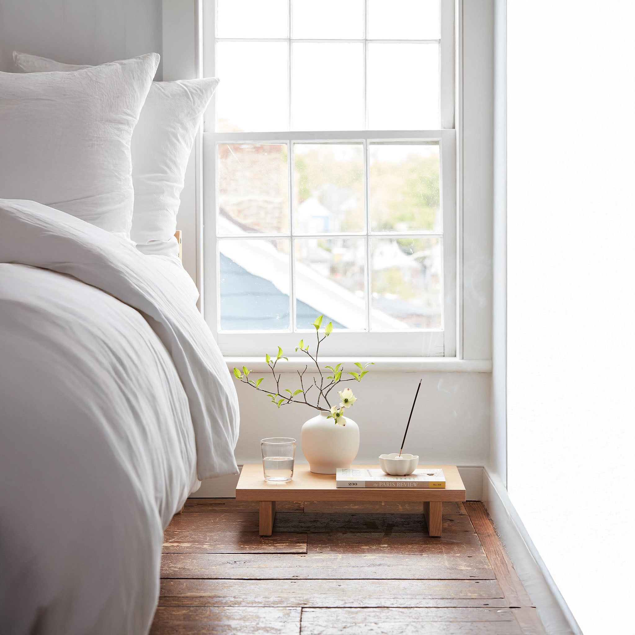 Serene bedside scene with a white ceramic fleur incense holder and minimalist ceramic vase on a low wooden platform, styled with a branch of spring blossoms and a lit incense stick.
