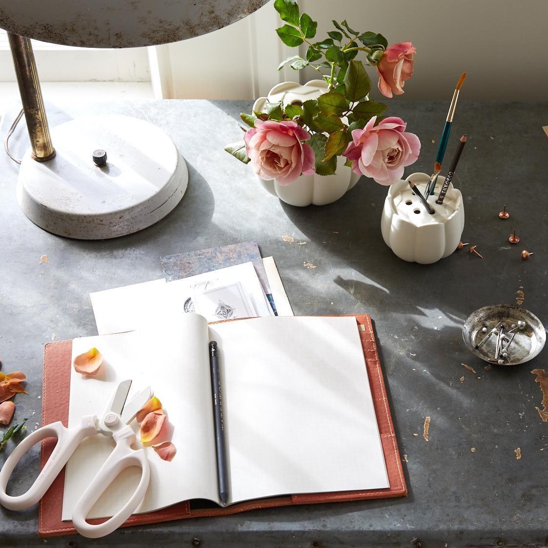 Bright tabletop scene featuring a ceramic flower frog vase holding pink garden roses, a matching ceramic holder with brushes and pencils, and a pair of white Japanese pruning scissors resting on an open notebook.