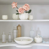 Shelf display featuring small and large white ceramic frog vases, styled with blush roses and surrounded by neutral-toned glass bottles and vessels.