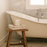 Bathroom with a bathtub, wooden stool, books and a hand poured vegan candle stacked on top.
