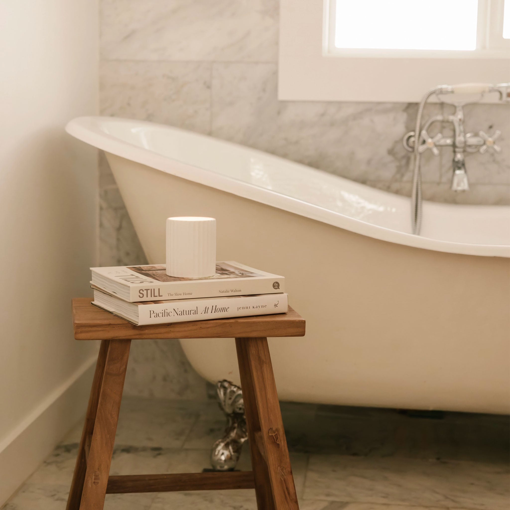 Bathroom with a bathtub, wooden stool, books and a hand poured vegan candle stacked on top.