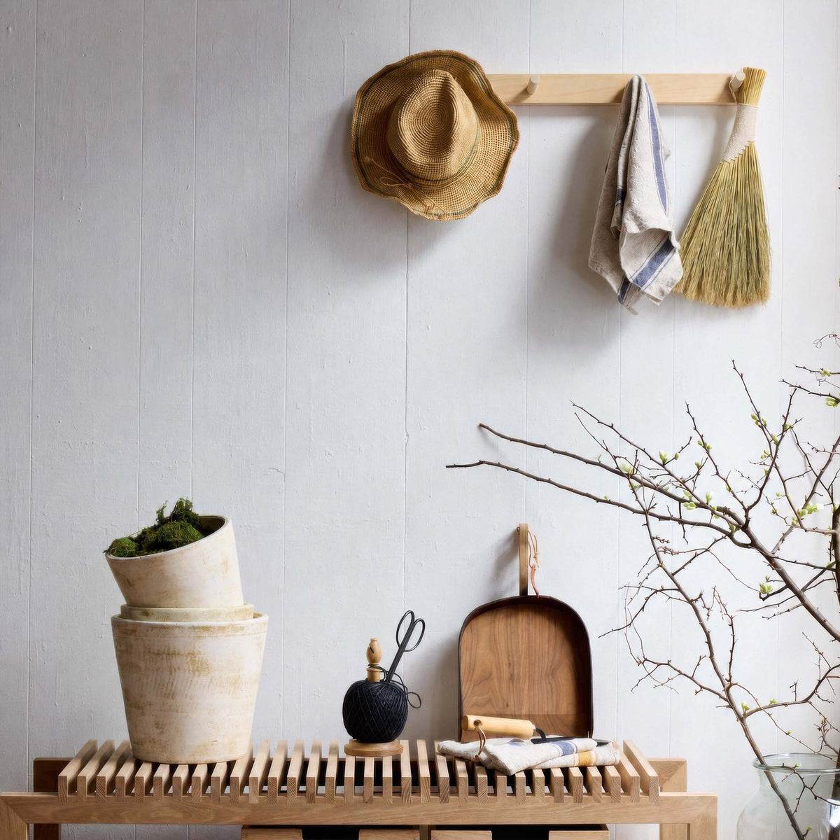 A light natural wood peg rack mounted on a white paneled wall, styled with a straw hat, a striped cloth, and a straw broom. Below, a matching wooden bench holds potted greenery, a small black bottle, a wooden scoop, and a cloth, with bare tree branches entering from the right.