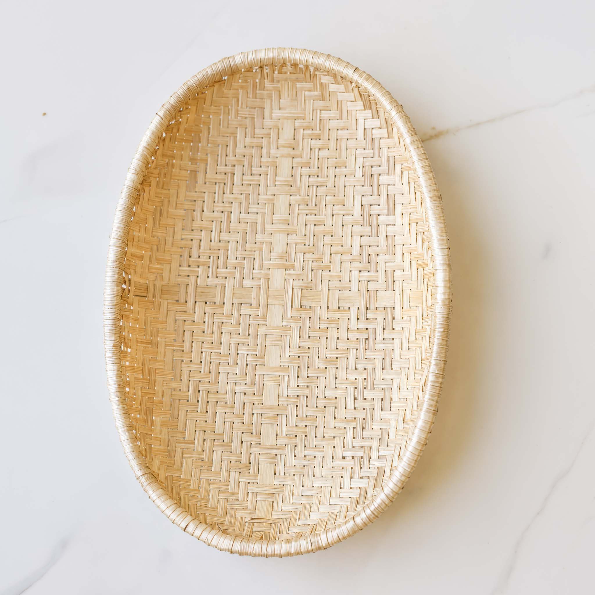 An oval woven tray made of natural light wood or rattan, photographed from above on a white marble surface.
