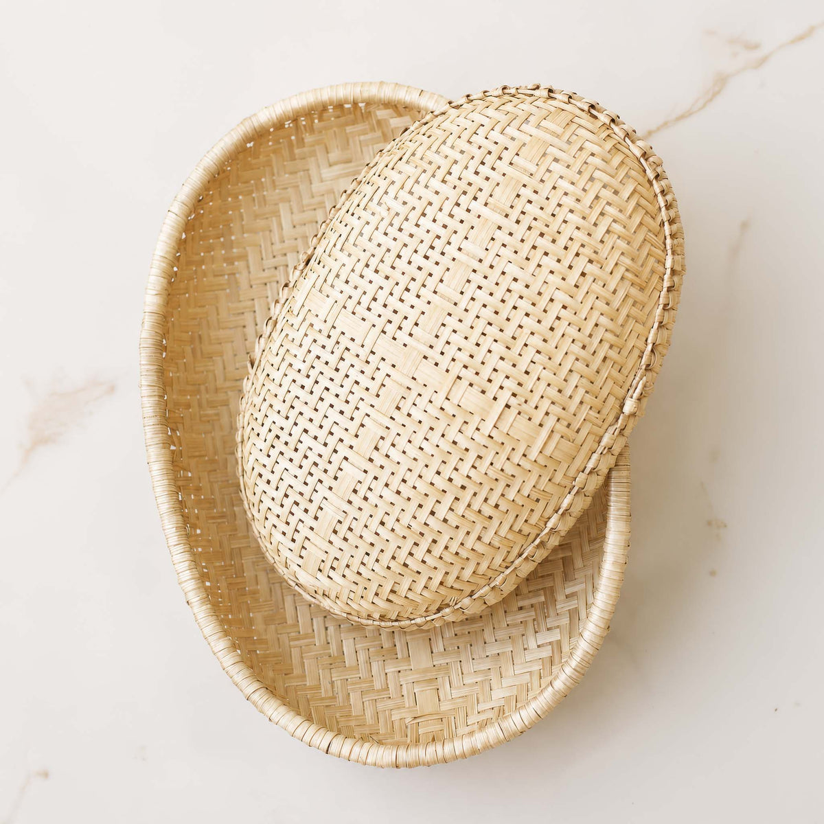 Two woven oval baskets one smaller, one larger, photographed from above on a light marble-like surface.