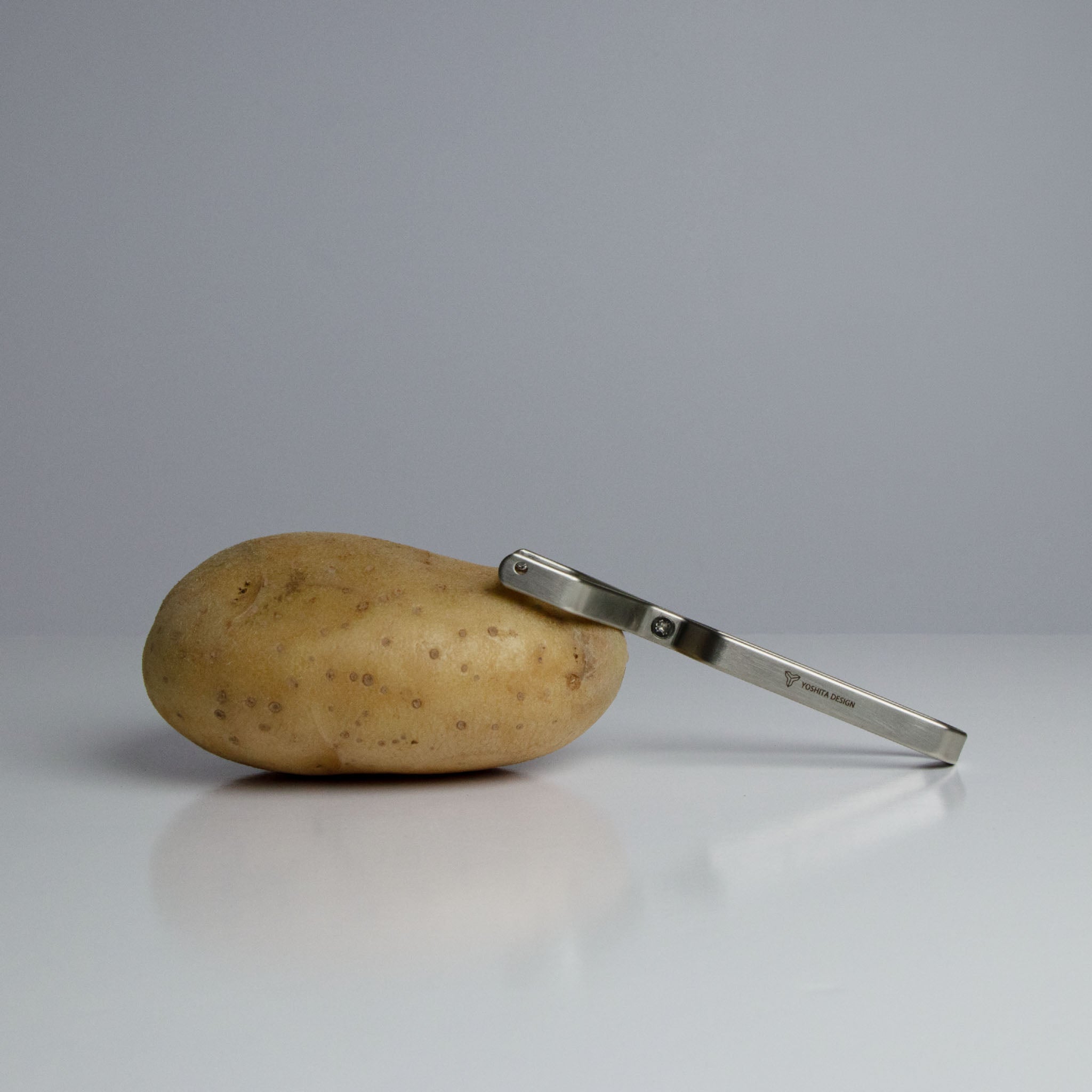 A potato and the Yoshital stainless steel peeler on a gray background