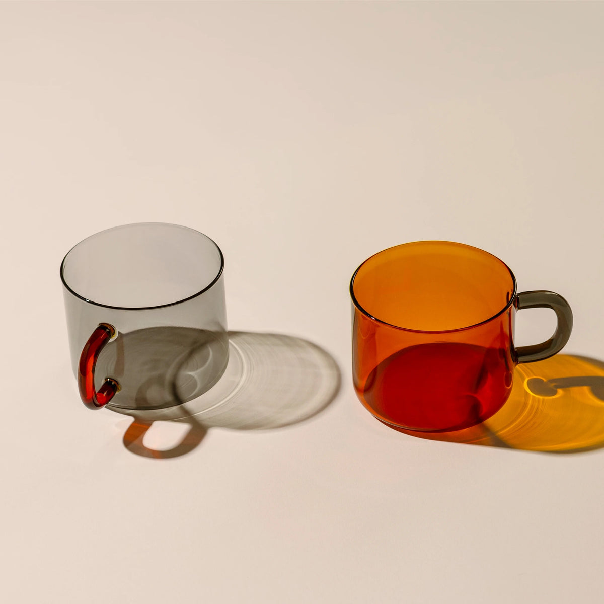 Two translucent glass mugs with thick handles on a light cream background. The mug on the left is amber and has a dark gray handle, while the mug on the right is gray with an orange handle.