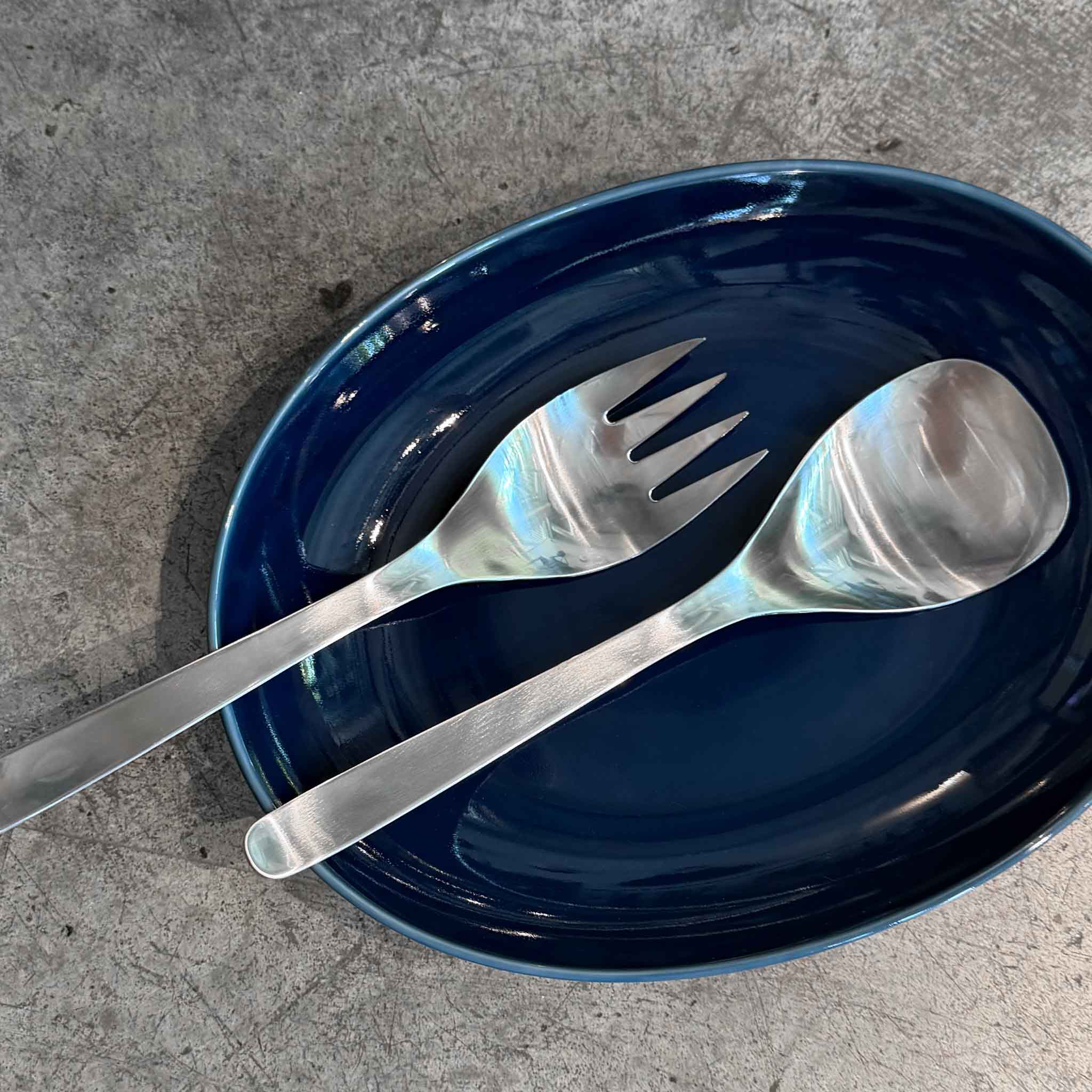 A stainless steel set of serving spoon and fork resting on a glossy dark blue oval plate, set on a textured gray concrete surface.