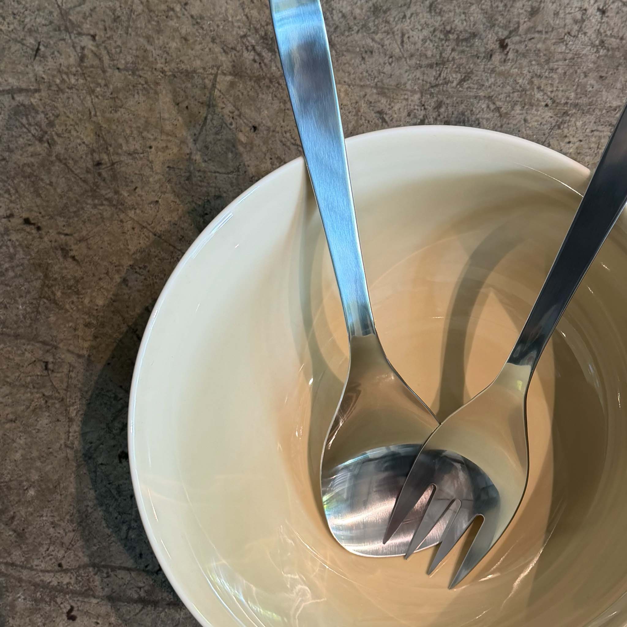 Two stainless steel utensils, a spoon and a fork, resting inside a shallow cream-colored ceramic bowl on a textured gray stone surface.