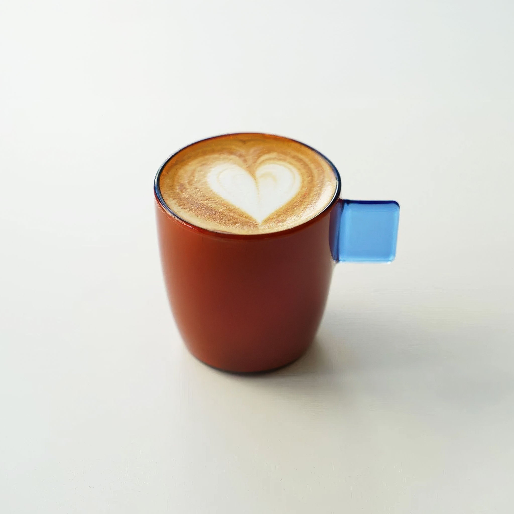Amber mug with blue handle containing a cappuccino with a heart design on a white background