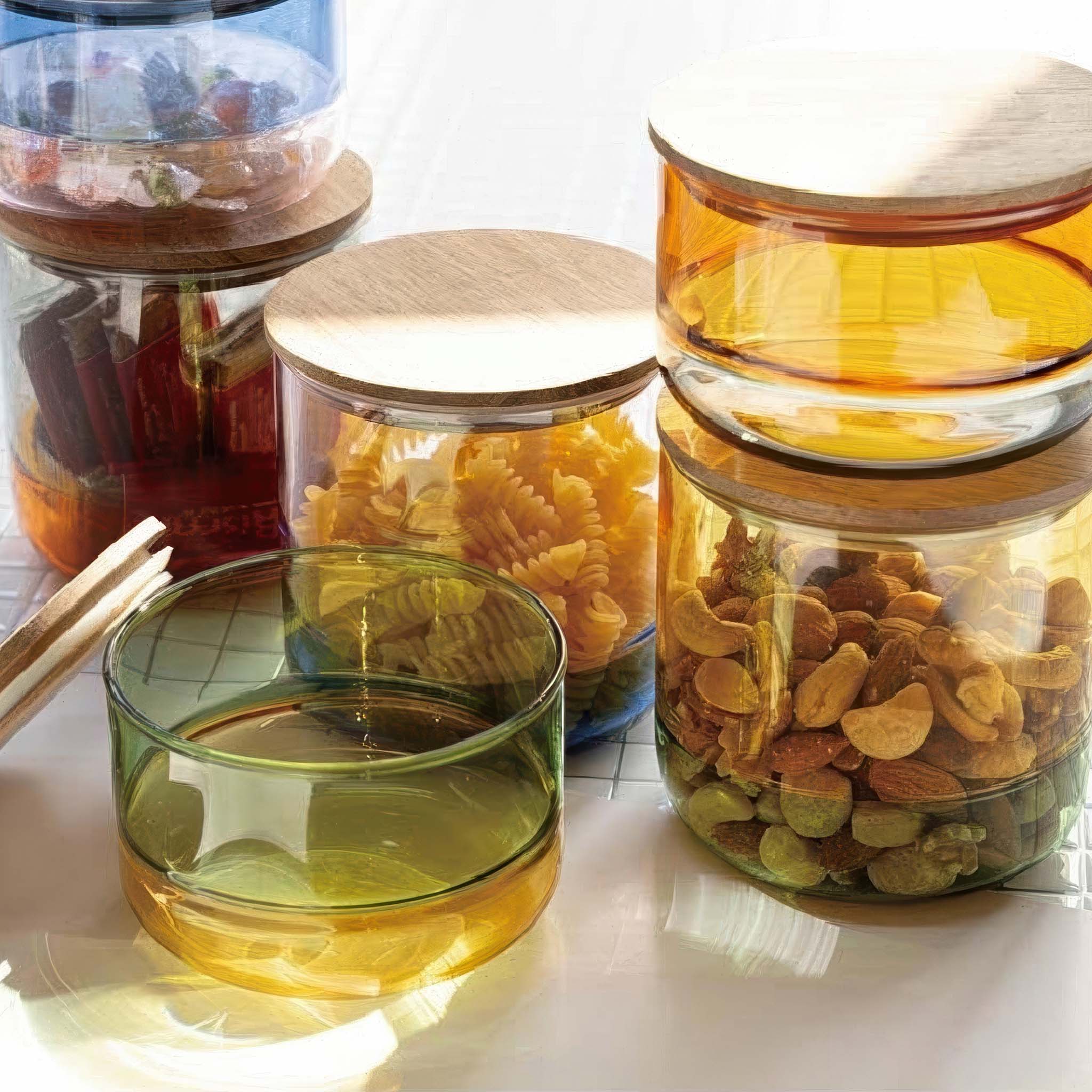 Set of Amabro glass canisters with wooden lids containing various snacks on a white surface.