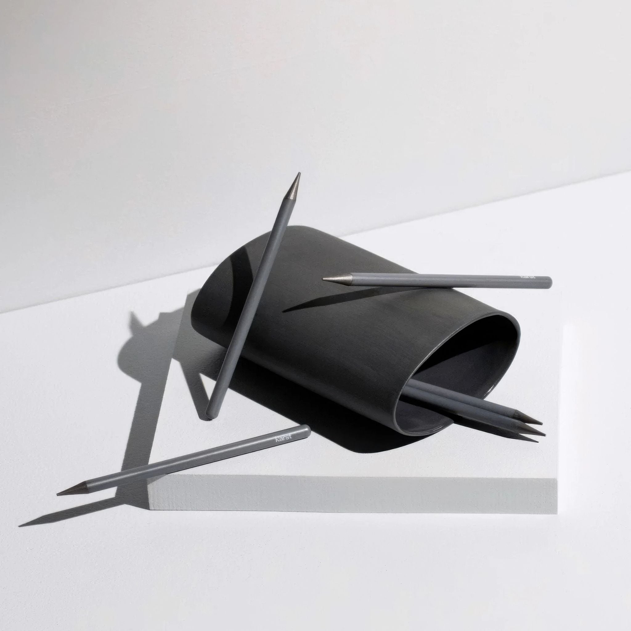 Karst woodless graphite pencils with matte finish, scattered around a modern black display case on a minimal white backdrop.