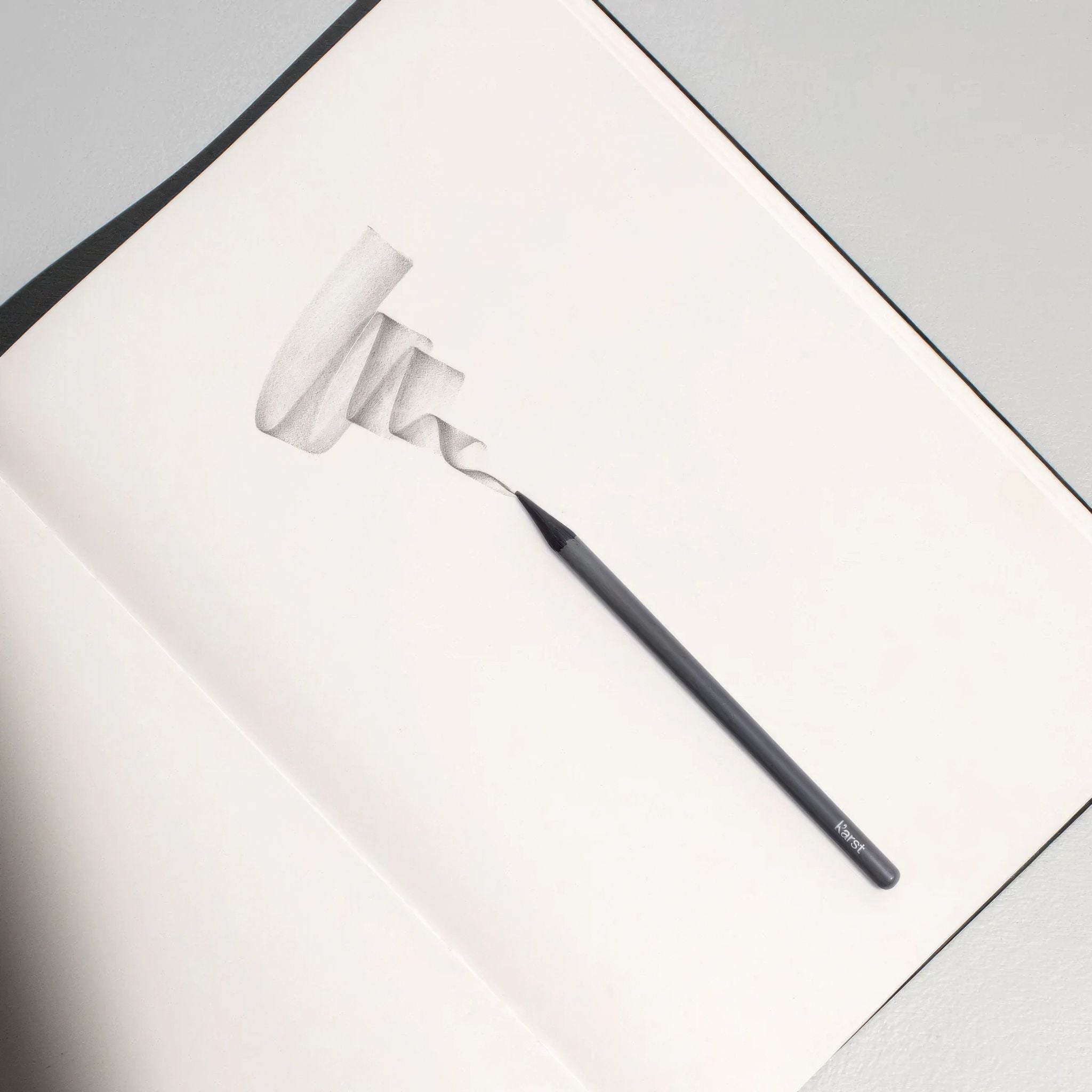 Karst woodless graphite pencil sketching a shaded ribbon design on white paper.