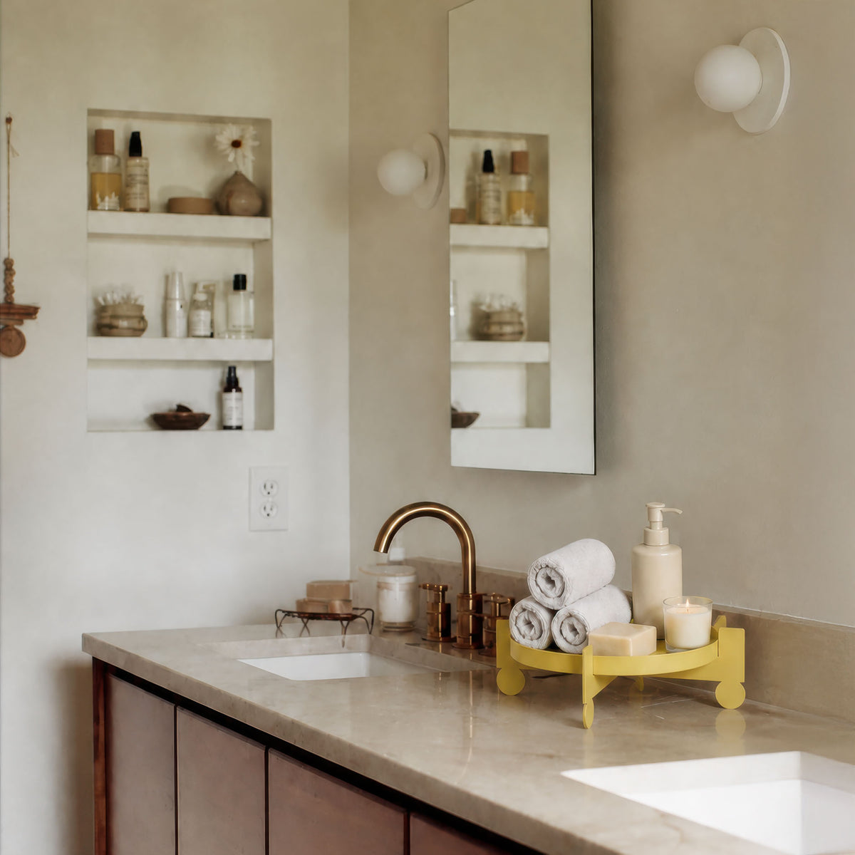A styled bathroom vanity scene with a beige countertop, white sinks, a brass faucet, built-in wall shelves, a mirror, and a yellow roll-bar tray holding rolled towels, soap bars, and a pump bottle.