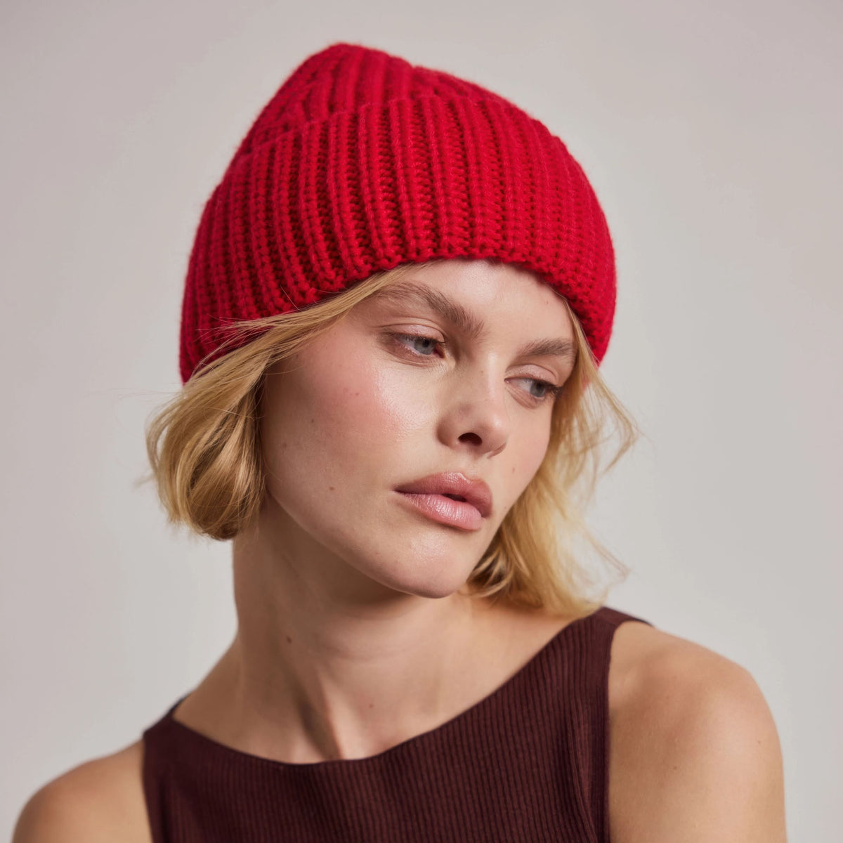 A close-up of a woman wearing a bright red ribbed knit beanie hat, paired with a dark brown ribbed top, against a neutral light gray background.