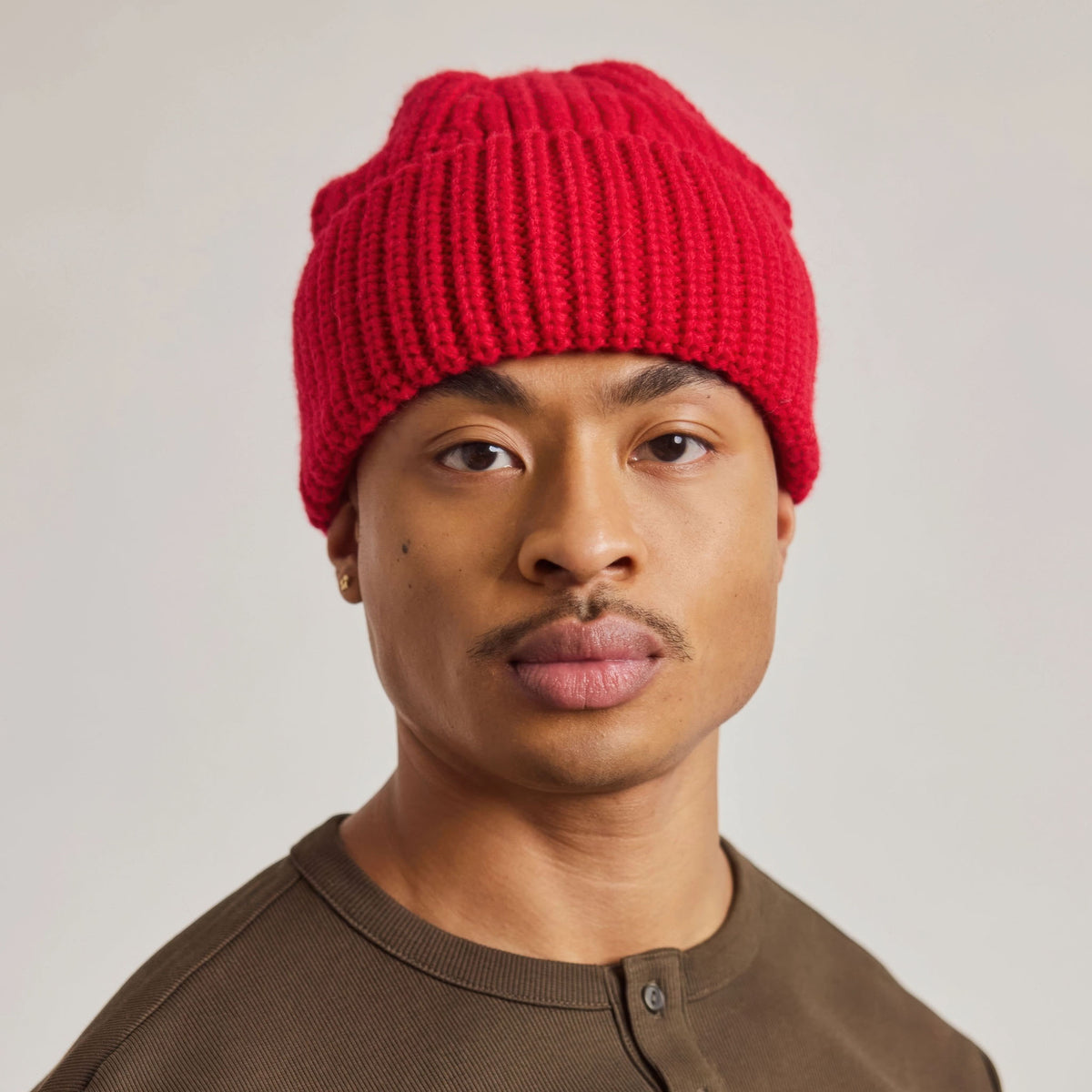 A man wearing a bright red ribbed knit beanie hat and a brown henley shirt, photographed against a plain light gray background.