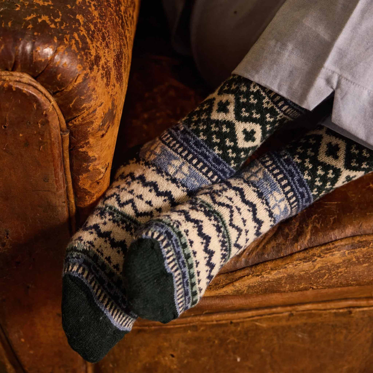 A close-up of a person's feet wearing lisa b's Fair Isle knit socks in Ivy, resting on a rustic brown leather armchair.