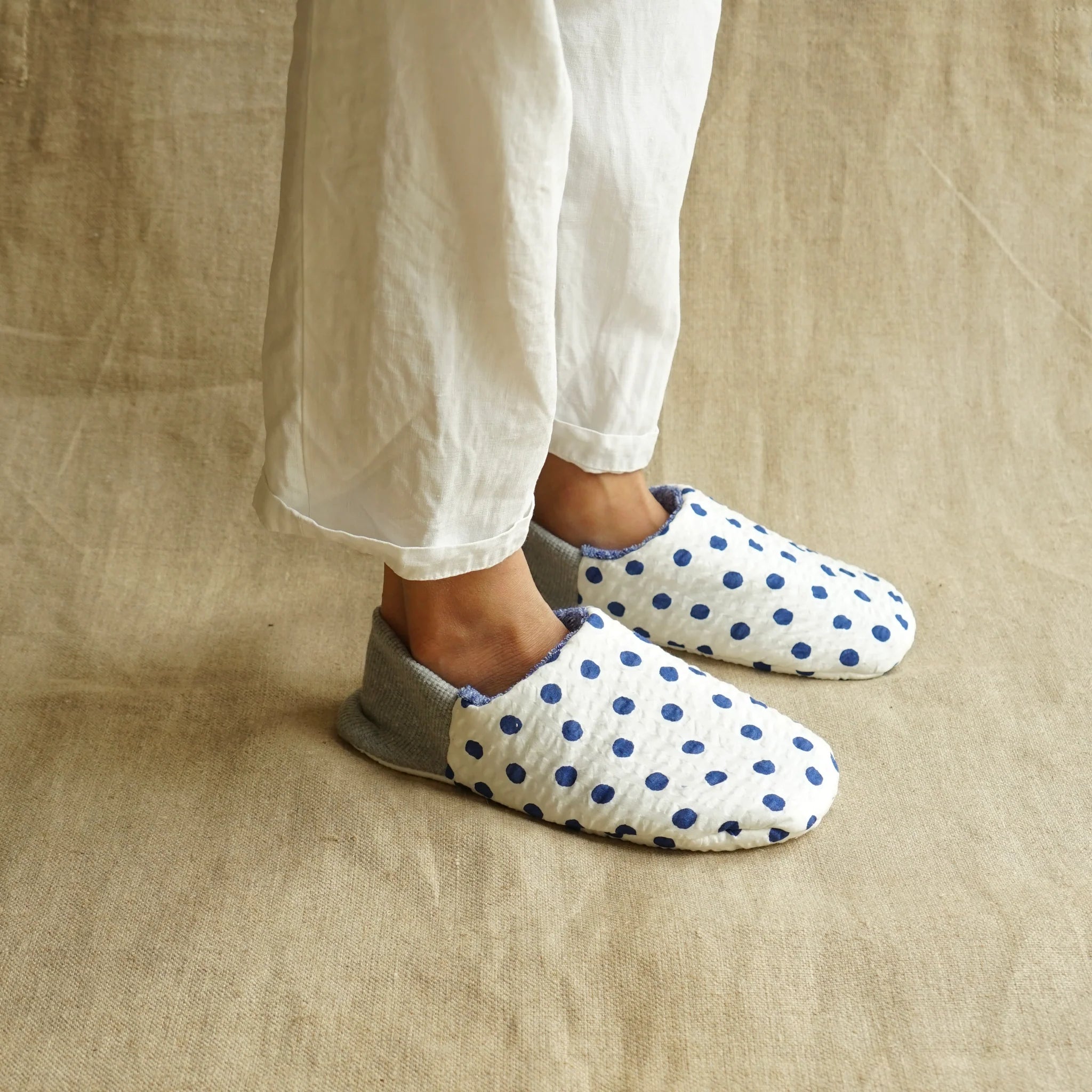 A person wearing white pants with blue and white polka dot slippers on a beige background