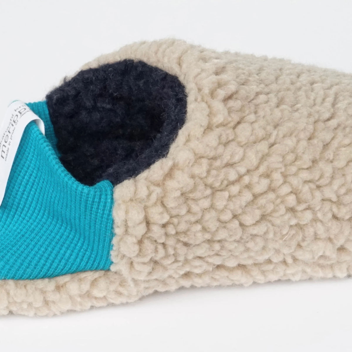 A close-up of a slip-on Merippa slipper with a chunky cream sherpa exterior, a bright turquoise ribbed knit collar, and a dark navy plush interior.