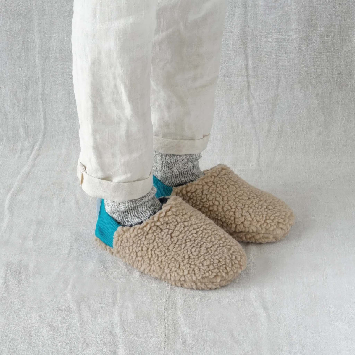 A person wearing Merippa beige sherpa fleece slip-on slippers with bright turquoise blue heel tabs, paired with gray socks and off-white trousers, standing against a white fabric backdrop.