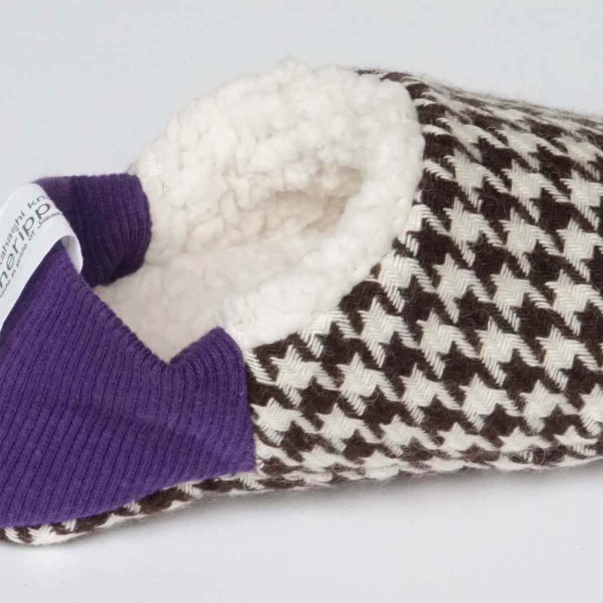 A close-up of a Merippa slip-on slipper with a brown and cream houndstooth pattern, a purple ribbed knit collar, and a white sherpa lining.