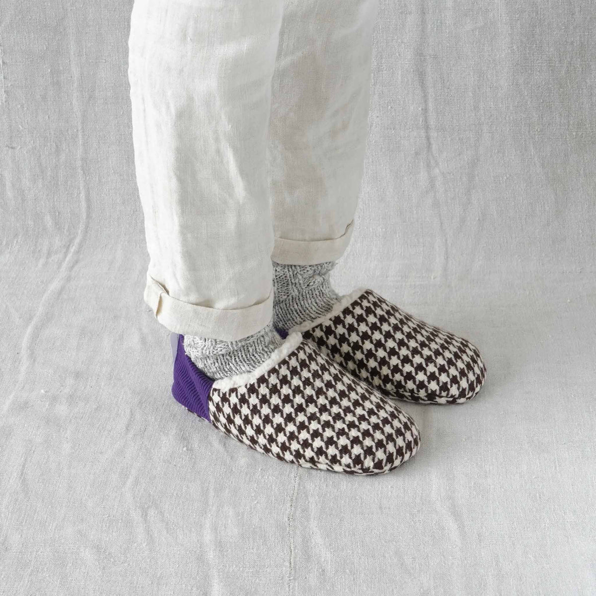 A person wearing white cuffed trousers, gray socks, and brown-and-white houndstooth Merippa slip-on slippers with a purple heel accent, standing against a light fabric backdrop.